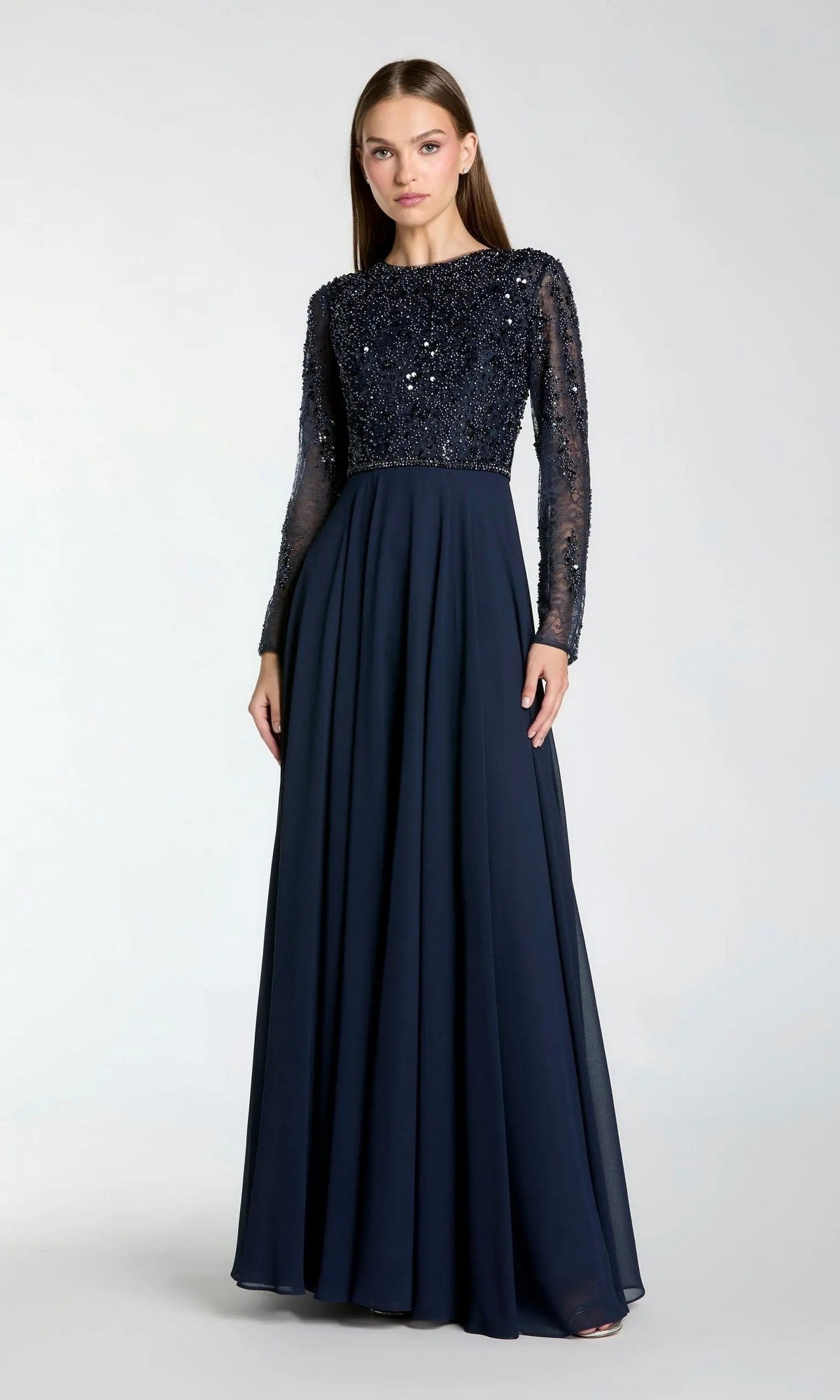 A woman wearing the Mac Duggal 21316 long formal dress in midnight, featuring an embellished sequined bodice and sheer long sleeves, stands against a plain light background.